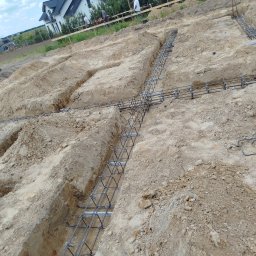 Viktor Kozachuk Uslugi OgulnoBudowlane - Zbrojenie fundamentów w wykopach, przygotowane do zalania betonem. Widoczne pręty zbrojeniowe połączone w siatkę. W tle budynki mieszkalne i ekipa budowlana.