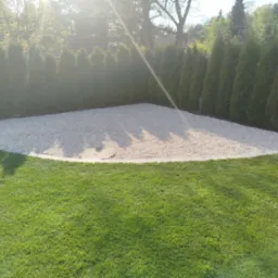 Ukończona aranżacja ogrodu: wysypany żwirkiem teren w kształcie trapezu, obrzeża z kostki, zielony trawnik i żywopłot z tui w tle, w słoneczny dzień.