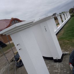 Viktor Kozachuk Uslugi OgulnoBudowlane - Biała, otynkowana zabudowa balkonu z widocznymi elementami mocującymi, na tle domu z czerwoną dachówką i pochmurnego nieba. Ujęcie z dołu, podkreślające perspektywę.