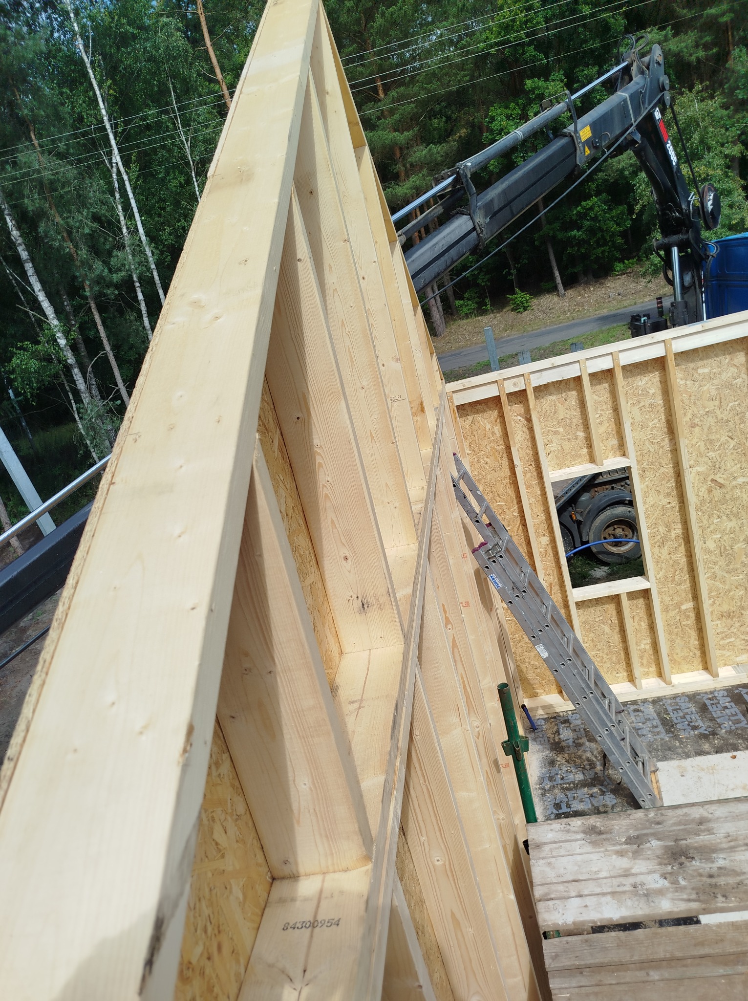 Konstrukcja drewniana ścian szkieletowych z widocznym oknem i dźwigiem w tle, etap budowy domu. Widok z góry na konstrukcję, z drabina opartą o ścianę.