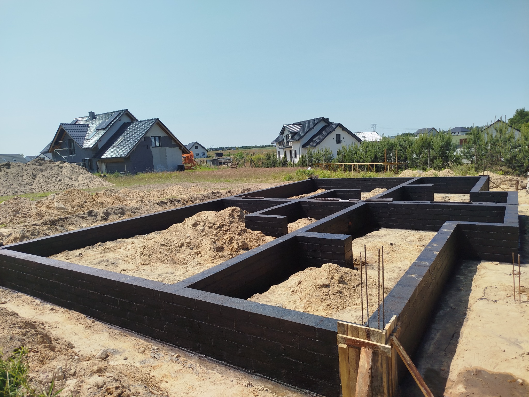 Fundamenty domu jednorodzinnego, czarne ściany z cegieł, widoczne zbrojenia i piasek wewnątrz, w tle zabudowa domów jednorodzinnych w słoneczny dzień.