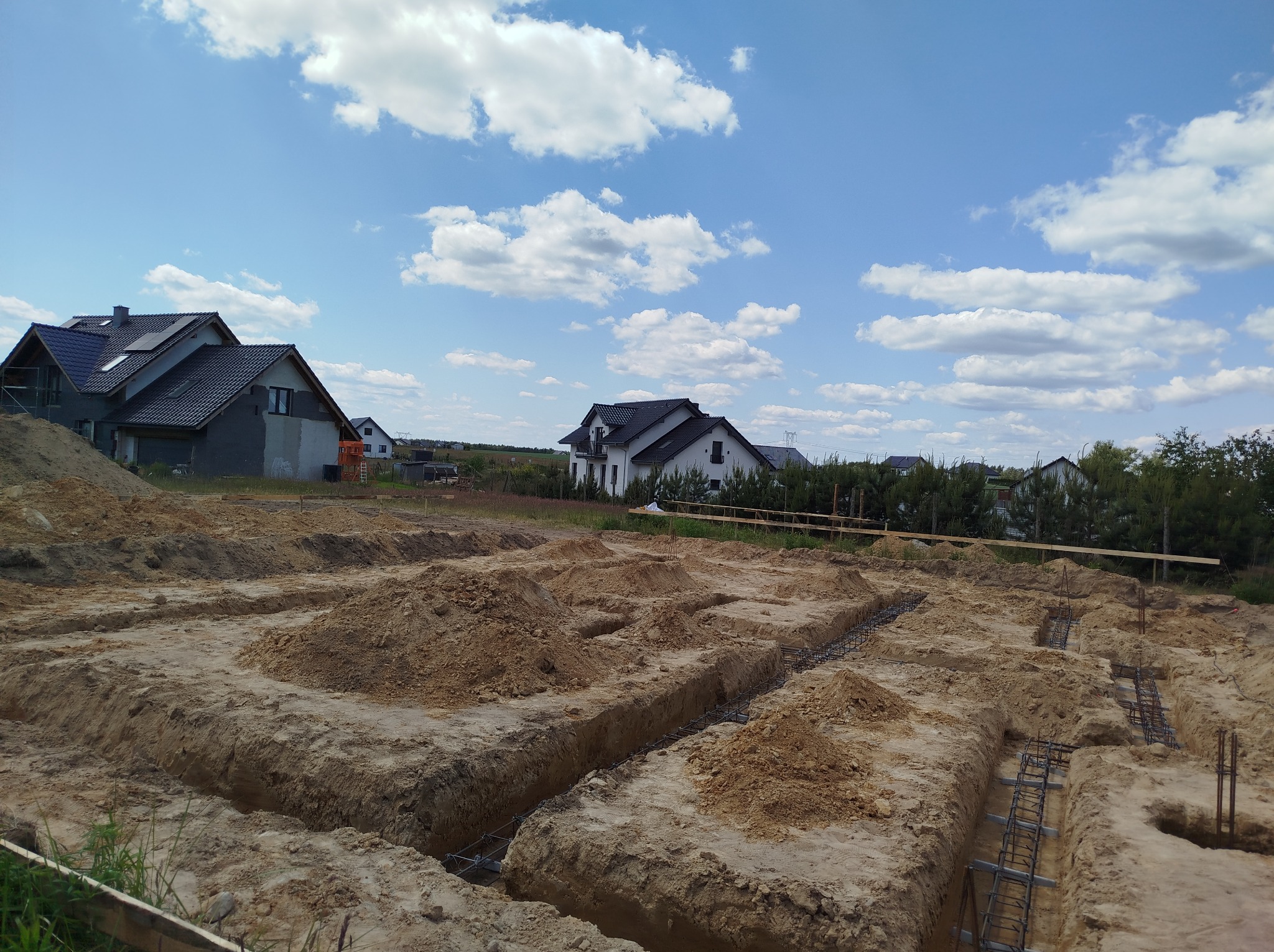 Wykonane wykopy pod fundamenty domu z widocznym zbrojeniem. Na dalszym planie widoczne budynki mieszkalne i niebieskie niebo z chmurami.