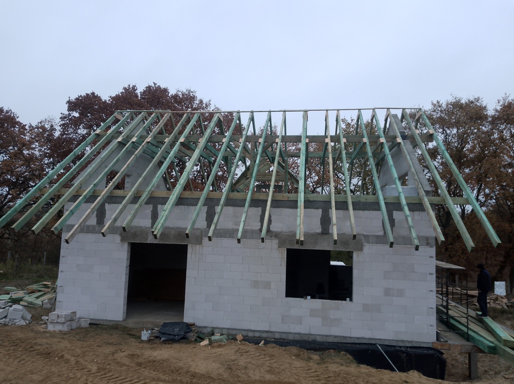 Wznoszenie domu z bloczków: surowy stan z drewnianą konstrukcją dachu w trakcie budowy. Widoczne bloczki, drewno i osoba na zewnątrz budynku.