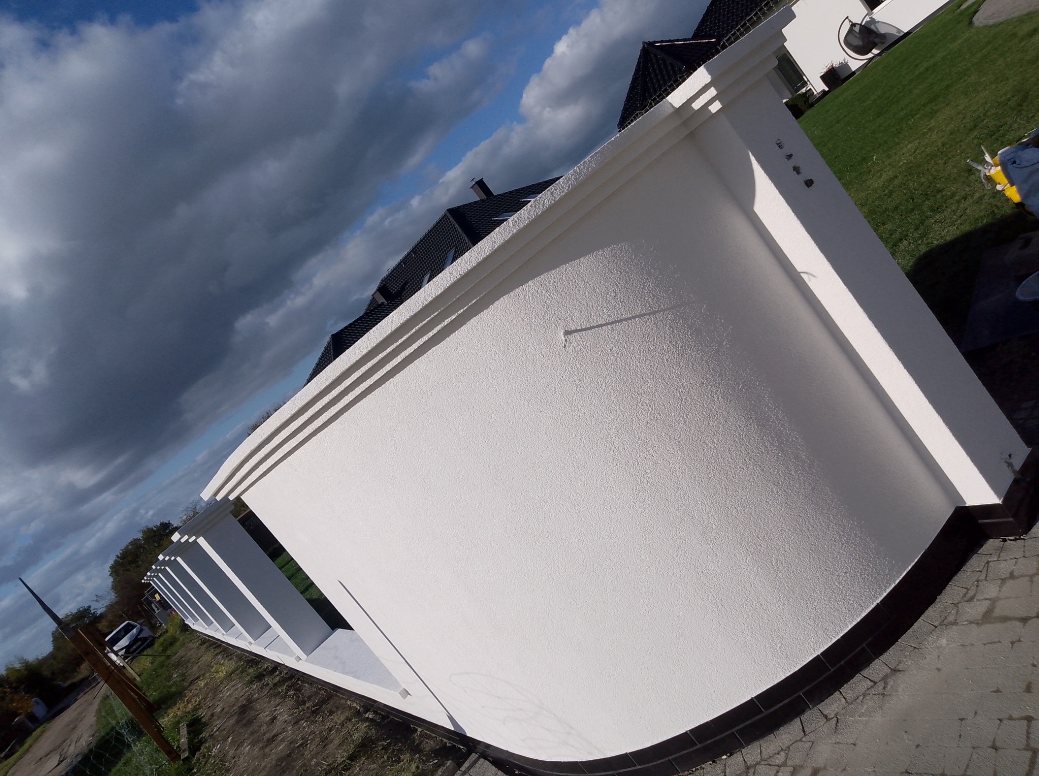 Biała, otynkowana ściana zabudowy balkonu z dekoracyjnymi elementami wzdłuż krawędzi, na tle pochmurnego nieba i fragmentu posesji.