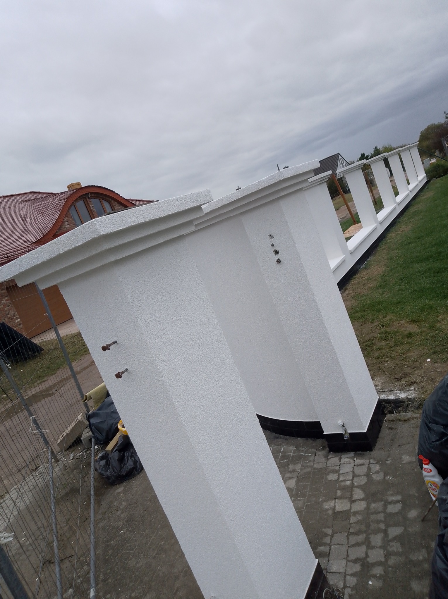 Biała, otynkowana zabudowa balkonu z widocznymi elementami mocującymi, na tle domu z czerwoną dachówką i pochmurnego nieba. Ujęcie z dołu, podkreślające perspektywę.