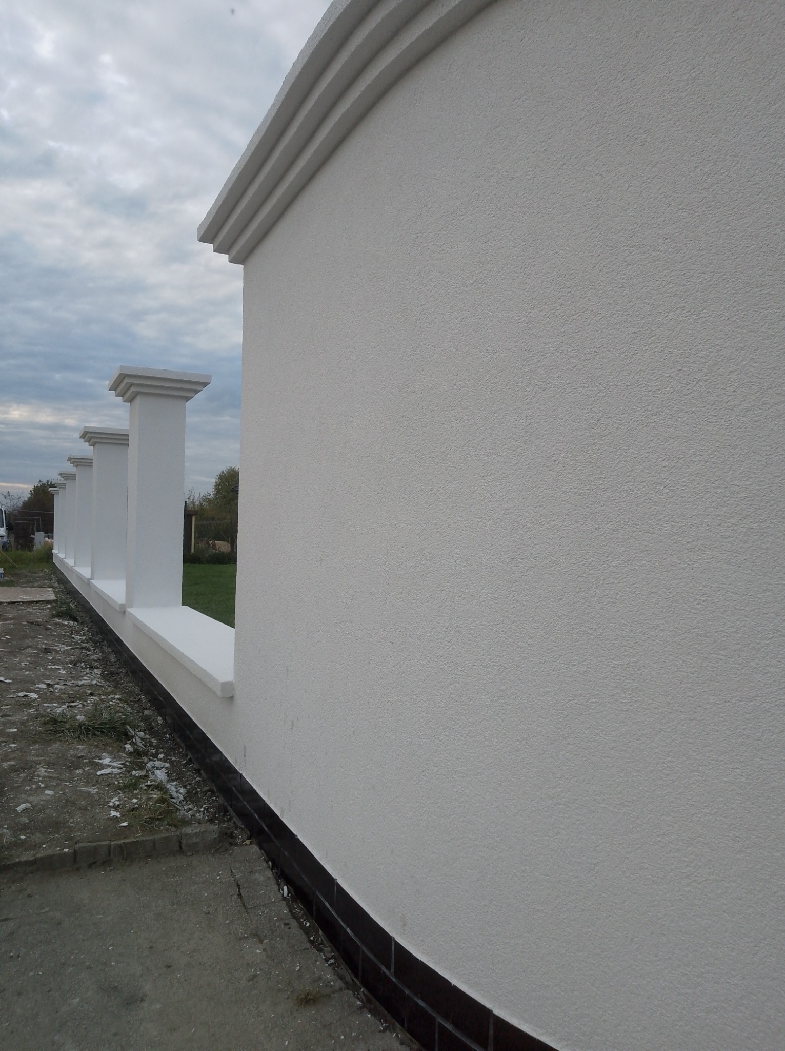 Wykończony fragment ogrodzenia z białego tynku strukturalnego i dekoracyjnymi, białymi słupkami. Widok z boku, na tle pochmurnego nieba i zieleni.