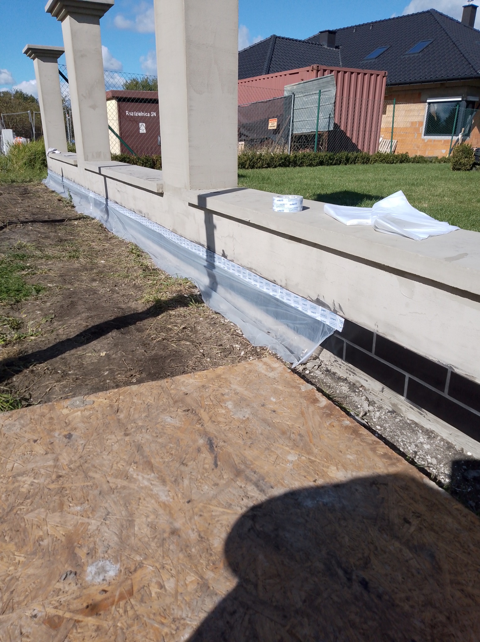 Przygotowanie do zabudowy balkonu z kolumnami, zabezpieczenie folią fundamentu. Widoczna taśma i arkusze folii ochronnej, płyta OSB na pierwszym planie.