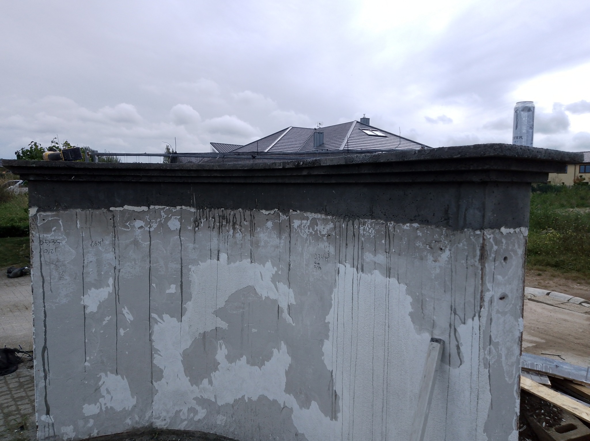 Szpachlowana ściana balkonu z widocznymi śladami po naprawach i notatkami, z fragmentem dachu domu w tle, sfotografowana w pochmurny dzień.