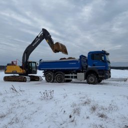 KOPALNIA MORYŃ Sp. z o.o. - Żółto-czarna koparka Volvo załadowuje urobek do niebieskiej wywrotki KH-Kipper na zasypanym śniegiem terenie. W tle widoczny las i pochmurne niebo.