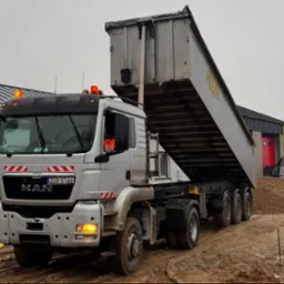 Ciężarówka MAN z podniesioną skrzynią ładunkową wysypuje żwir na plac budowy. W tle budynek z czerwoną bramą i zaparkowany bus.