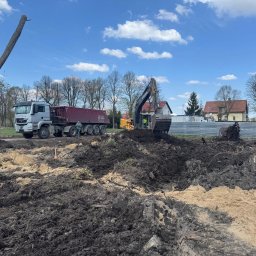 KOPALNIA MORYŃ Sp. z o.o. - Ciężarówka MAN i koparka Volvo na placu budowy. W tle domy i błękitne niebo z chmurami. Wykopana ziemia zmieszana z piaskiem, pozostałości po wycince drzew.