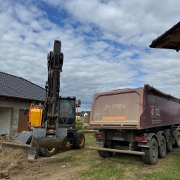 KOPALNIA MORYŃ Sp. z o.o. - Koparka kołowa z podniesionym ramieniem i wywrotka ZS 270RA na trawiastym terenie budowy, w tle dom z grafitowym dachem i pochmurne niebo.