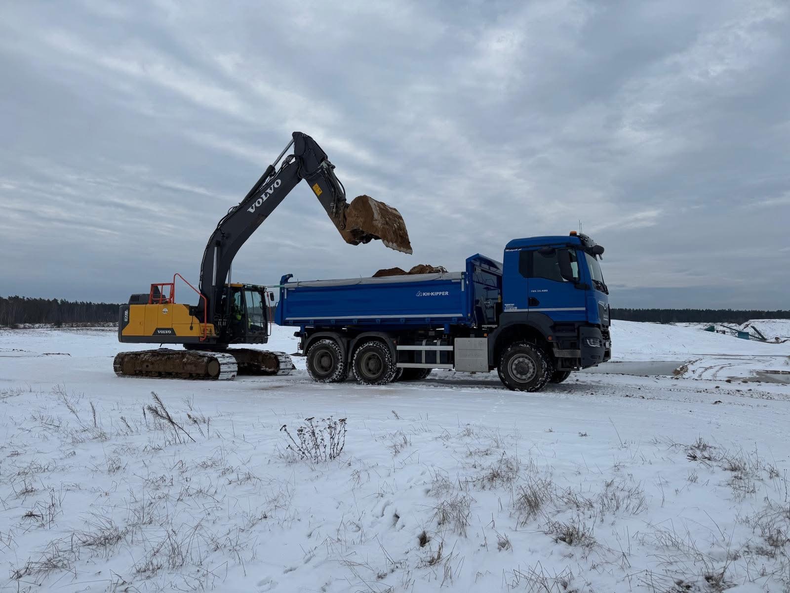 Żółto-czarna koparka Volvo załadowuje urobek do niebieskiej wywrotki KH-Kipper na zasypanym śniegiem terenie. W tle widoczny las i pochmurne niebo.