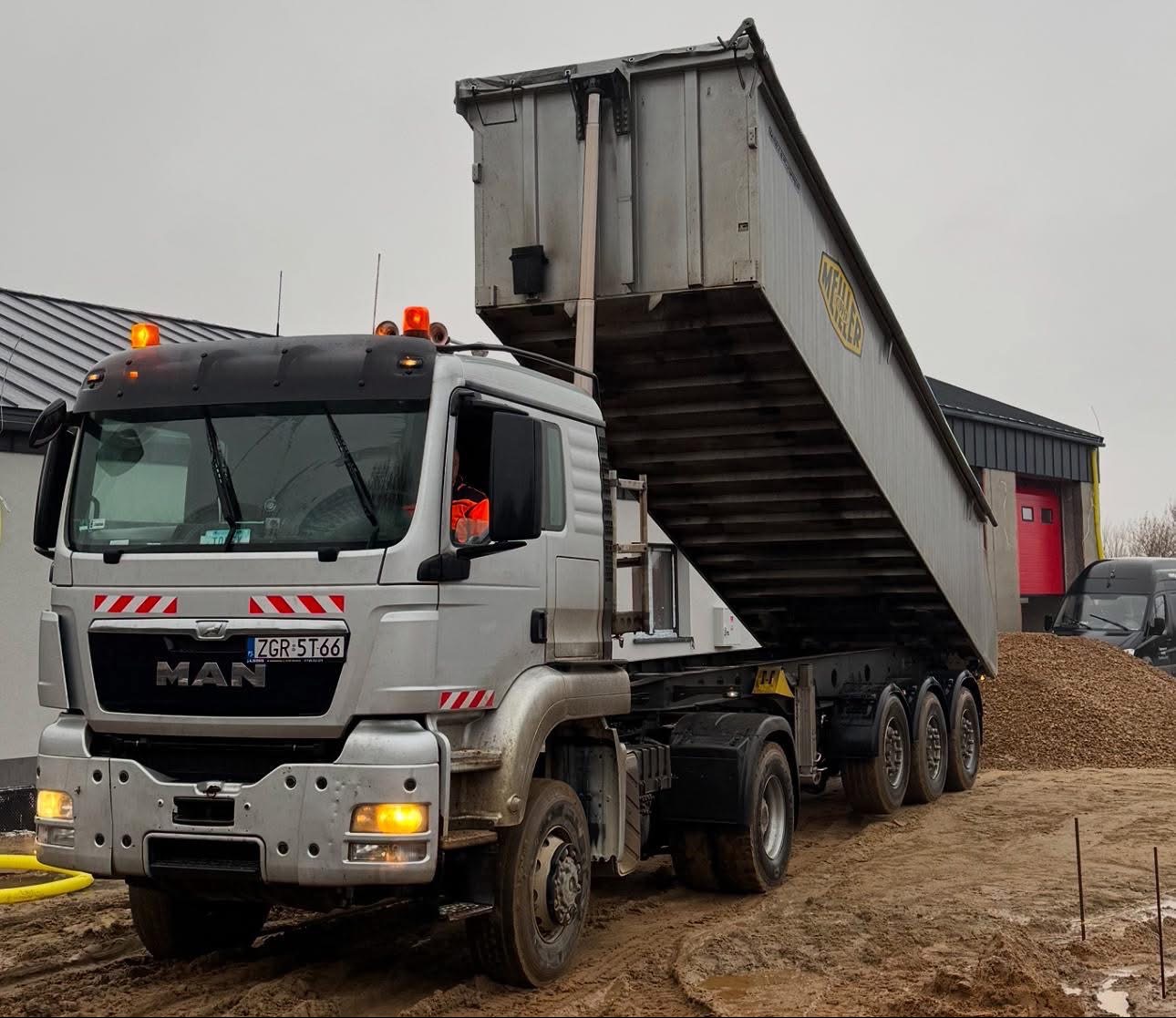 Ciężarówka MAN z podniesioną skrzynią ładunkową wysypuje żwir na plac budowy. W tle budynek z czerwoną bramą i zaparkowany bus.