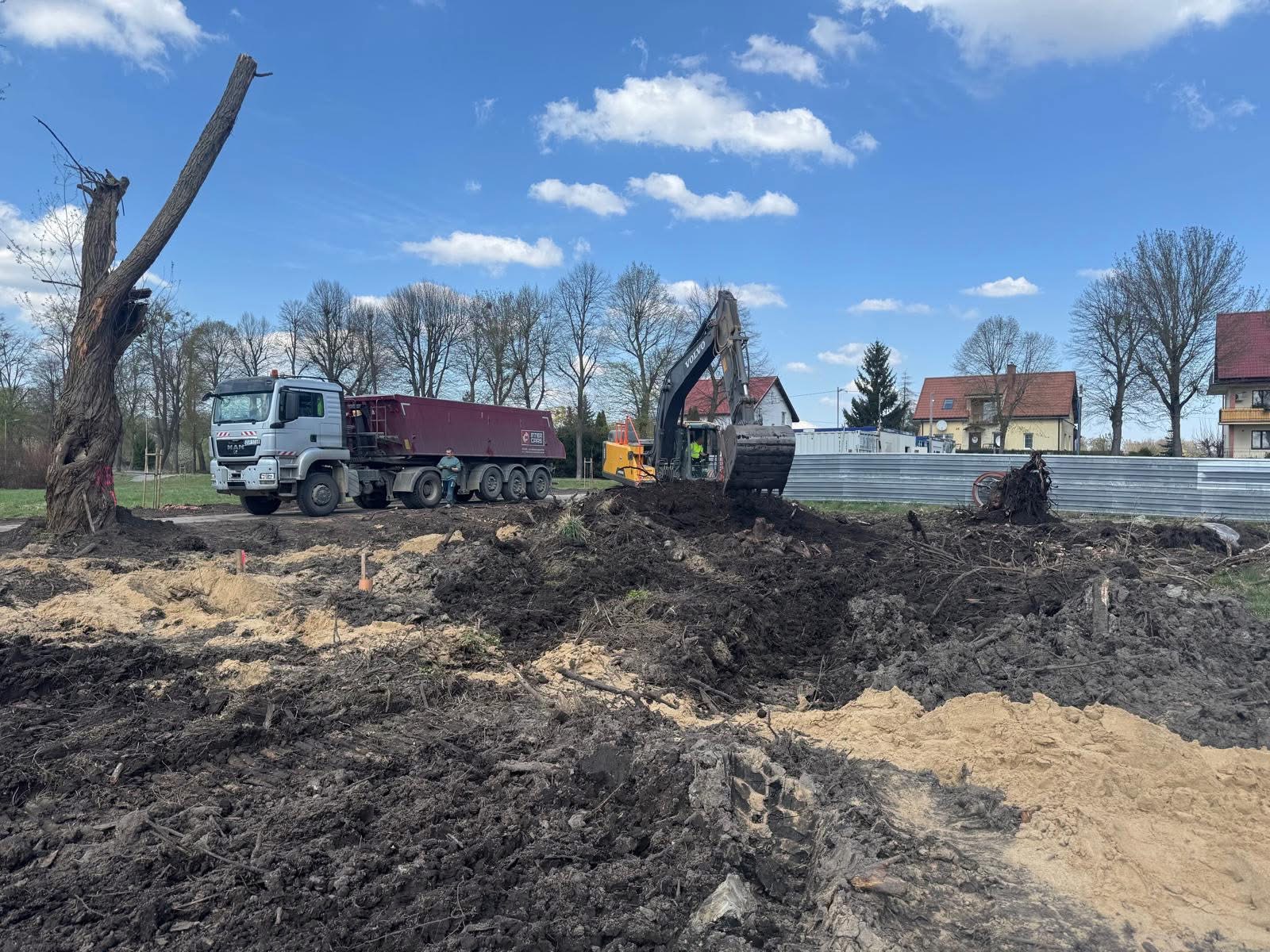 Ciężarówka MAN i koparka Volvo na placu budowy. W tle domy i błękitne niebo z chmurami. Wykopana ziemia zmieszana z piaskiem, pozostałości po wycince drzew.