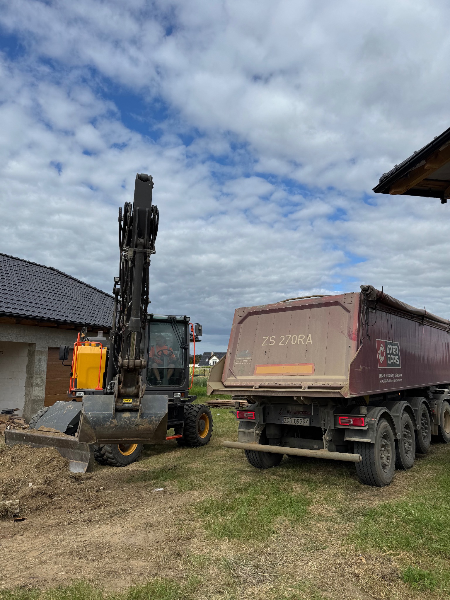 Koparka kołowa z podniesionym ramieniem i wywrotka ZS 270RA na trawiastym terenie budowy, w tle dom z grafitowym dachem i pochmurne niebo.