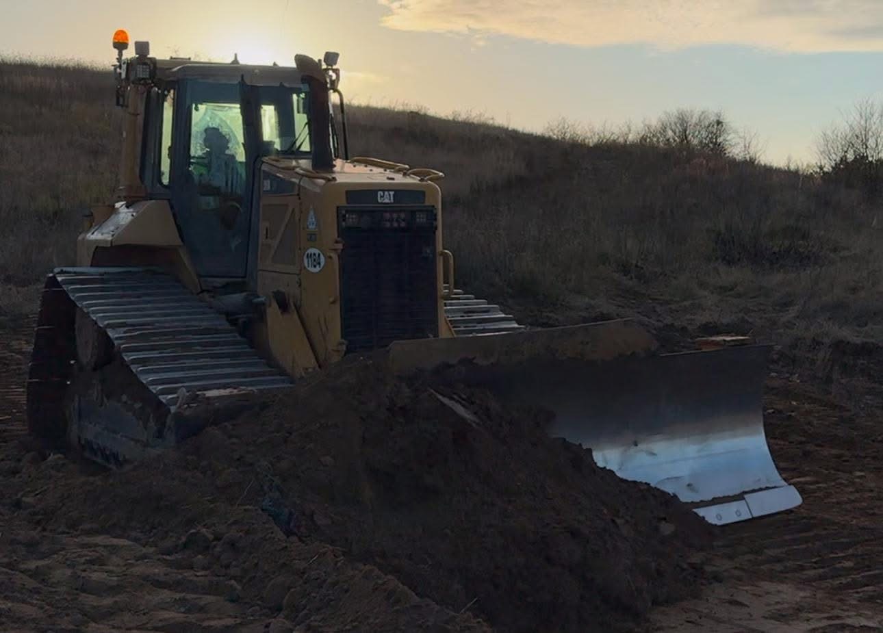 Żółty spychacz marki CAT podczas pracy na nierównym terenie, z widoczną stertą ziemi przed ostrzem i zachodzącym słońcem w tle.