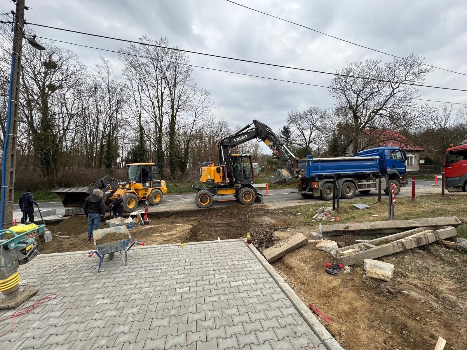 Układanie kostki brukowej z użyciem ciężkiego sprzętu Volvo: koparka i ładowarka. Widoczny fragment nowej nawierzchni i przygotowany teren. Praca wre.