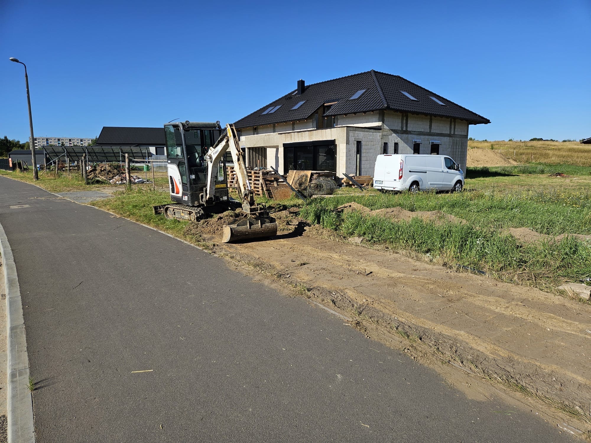 Mini koparka Bobcat przy wykopie obok drogi, w tle budynek w trakcie budowy z białym busem. Słoneczny dzień.