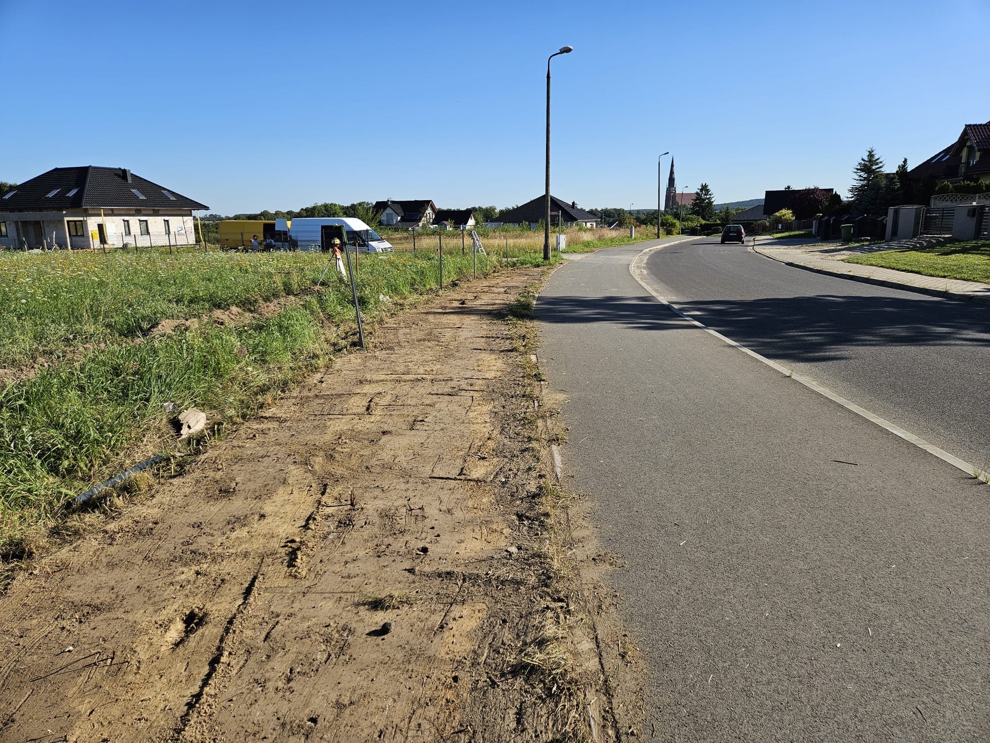 Przygotowany teren pod budowę, z widocznymi śladami po maszynach, graniczący z asfaltową drogą. W tle dom w trakcie budowy i zabudowa jednorodzinna.