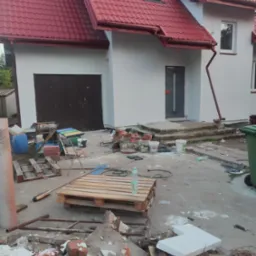 Dom jednorodzinny z czerwoną dachówką po remoncie elewacji, widoczne ślady prac budowlanych, palety, gruz, narzędzia i pojemniki na zewnątrz.