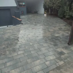 Ułożona, prostokątna kostka brukowa w odcieniach szarości i beżu na podjeździe przed domem z garażem, widoczne drzewa iglaste w tle. Powierzchnia kostki lekko mokra po deszczu.