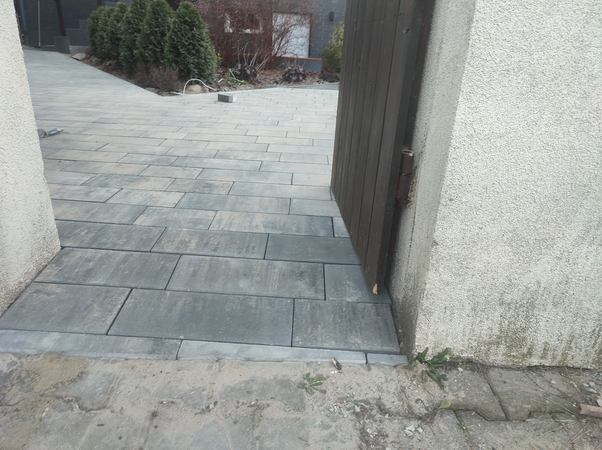 Nowa, szara kostka brukowa ułożona na podjeździe, widoczna brama wejściowa i fragment elewacji budynku z widocznymi śladami wilgoci.