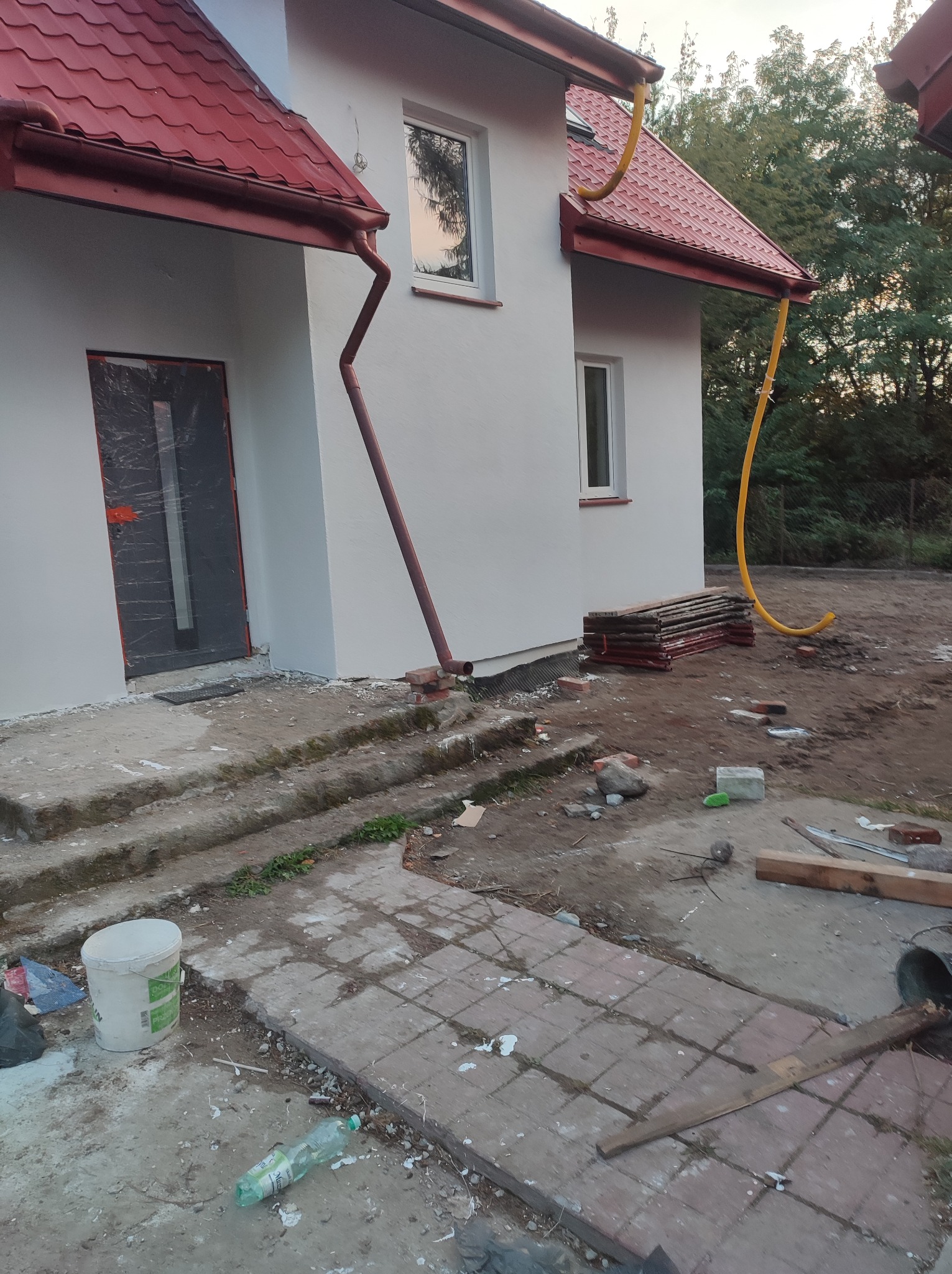 Świeżo pomalowana elewacja domu jednorodzinnego z nowymi rynnami, widoczne ślady prac budowlanych na podłożu i zabezpieczone folią drzwi wejściowe.