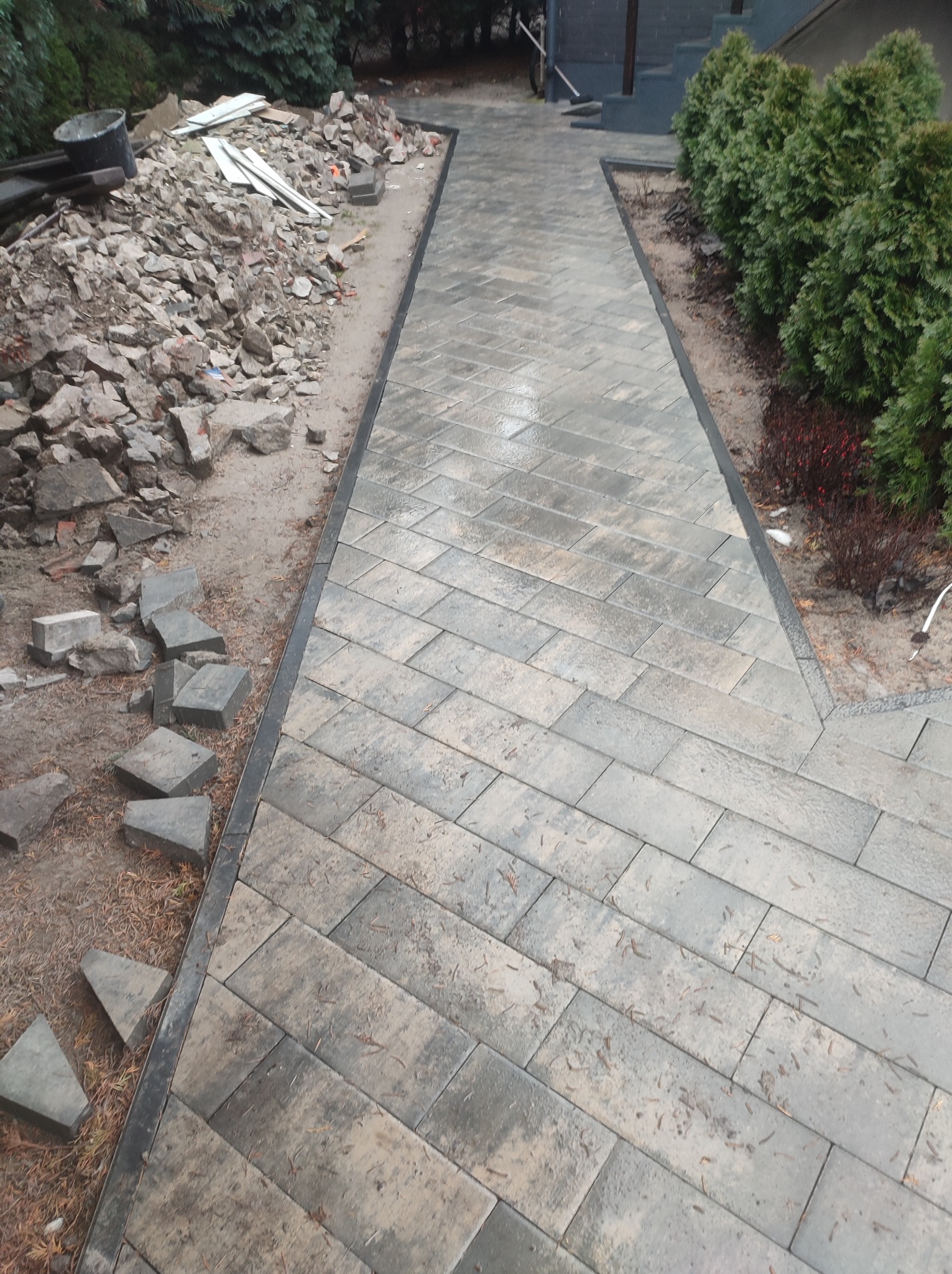Ułożona kostka brukowa wzdłuż ścieżki, z gruzem i przygotowanym podłożem w tle, widoczne tuje i schody wejściowe do budynku.