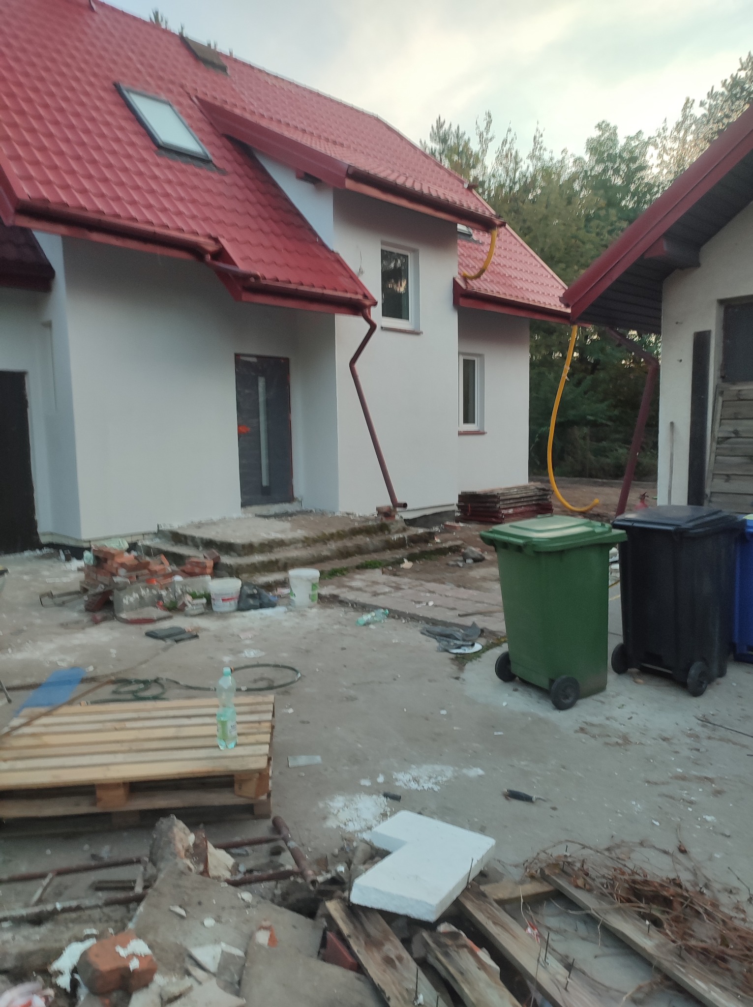 Dwukondygnacyjny dom z czerwoną dachówką w trakcie prac wykończeniowych, widoczne ślady po pracach budowlanych, gruz i pojemniki na śmieci.