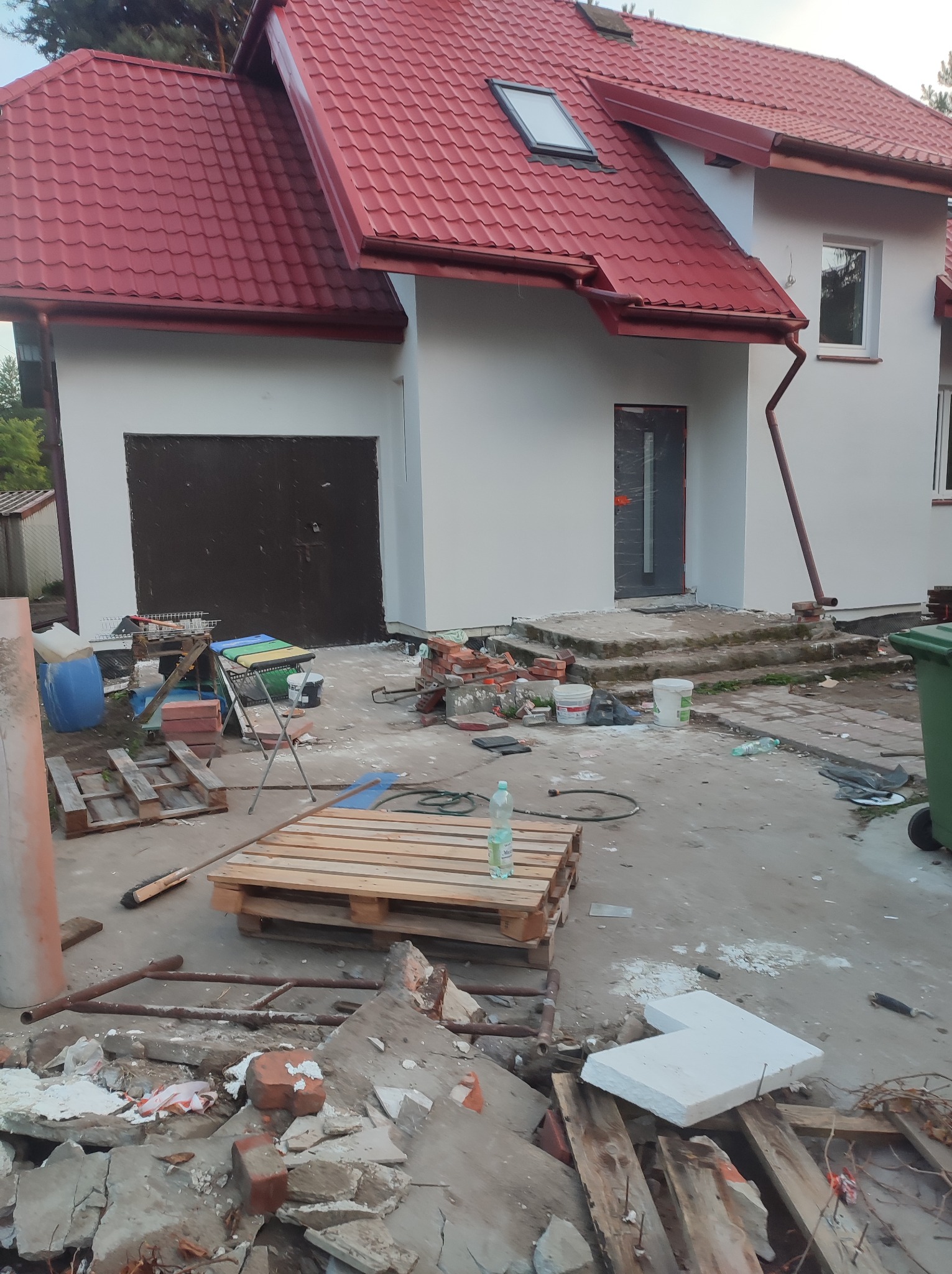 Dom jednorodzinny z czerwoną dachówką po remoncie elewacji, widoczne ślady prac budowlanych, palety, gruz, narzędzia i pojemniki na zewnątrz.