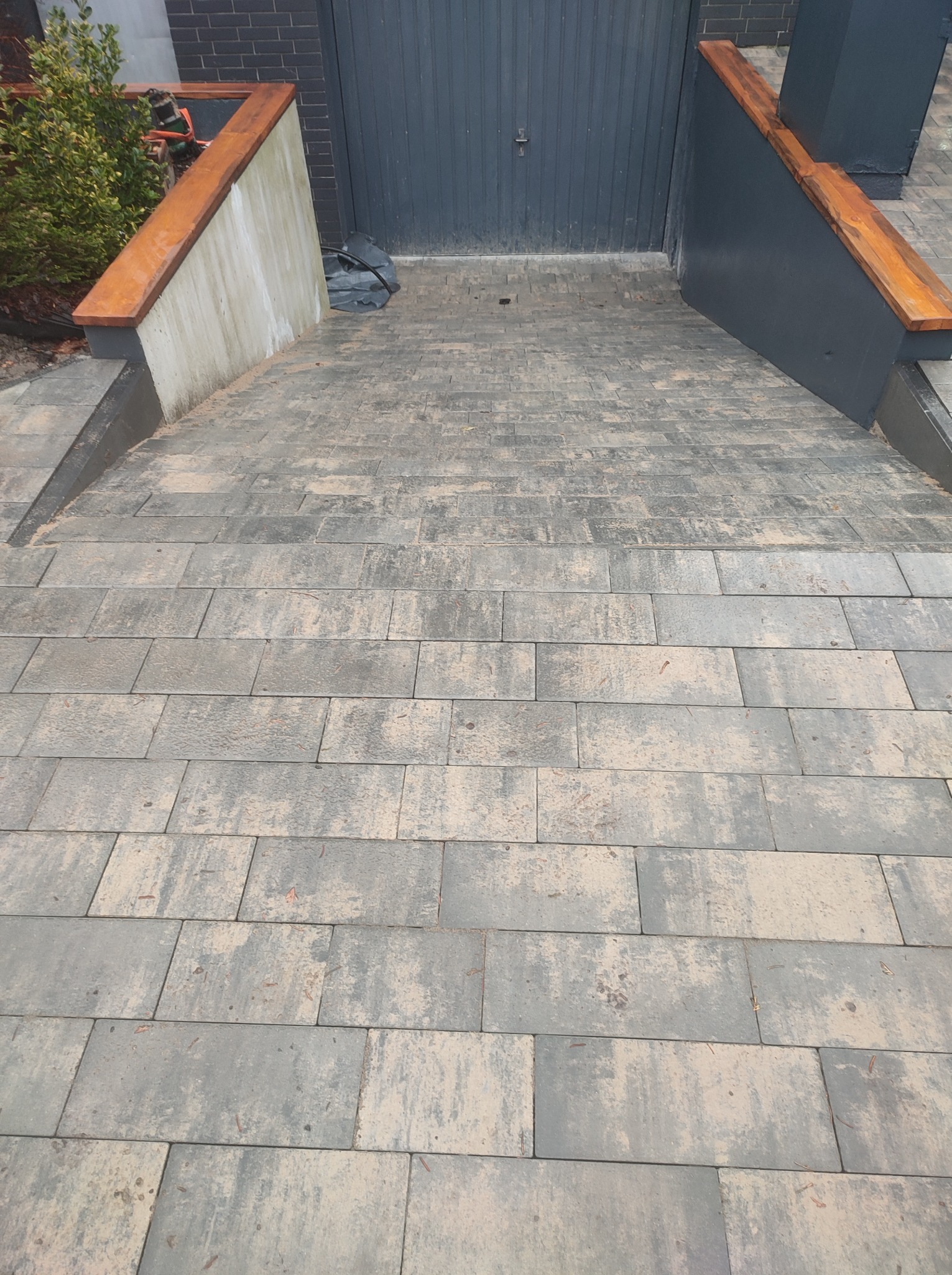 Szeroki najazd do garażu wyłożony szarą kostką brukową, zakończony niskimi ściankami z drewnianymi poręczami, widok z góry.