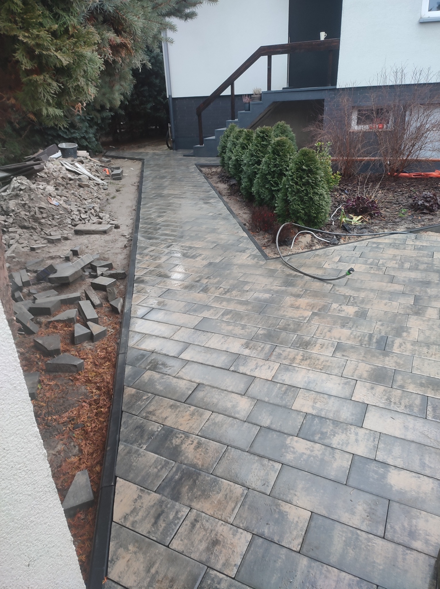 Układanie szarej kostki brukowej na ścieżce obok domu, widoczne nieułożone kostki i narzędzia budowlane.