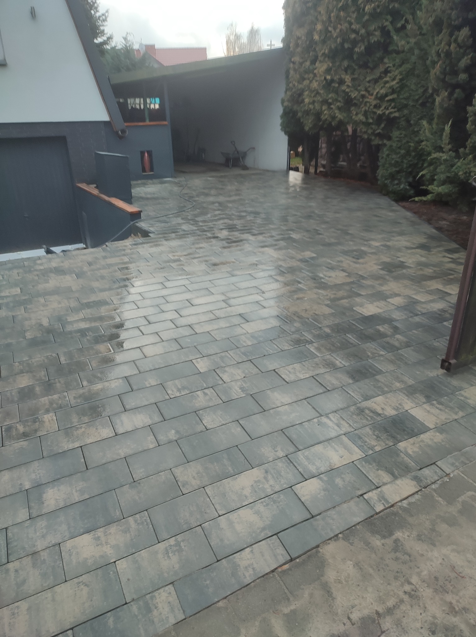 Ułożona, prostokątna kostka brukowa w odcieniach szarości i beżu na podjeździe przed domem z garażem, widoczne drzewa iglaste w tle. Powierzchnia kostki lekko mokra po deszczu.