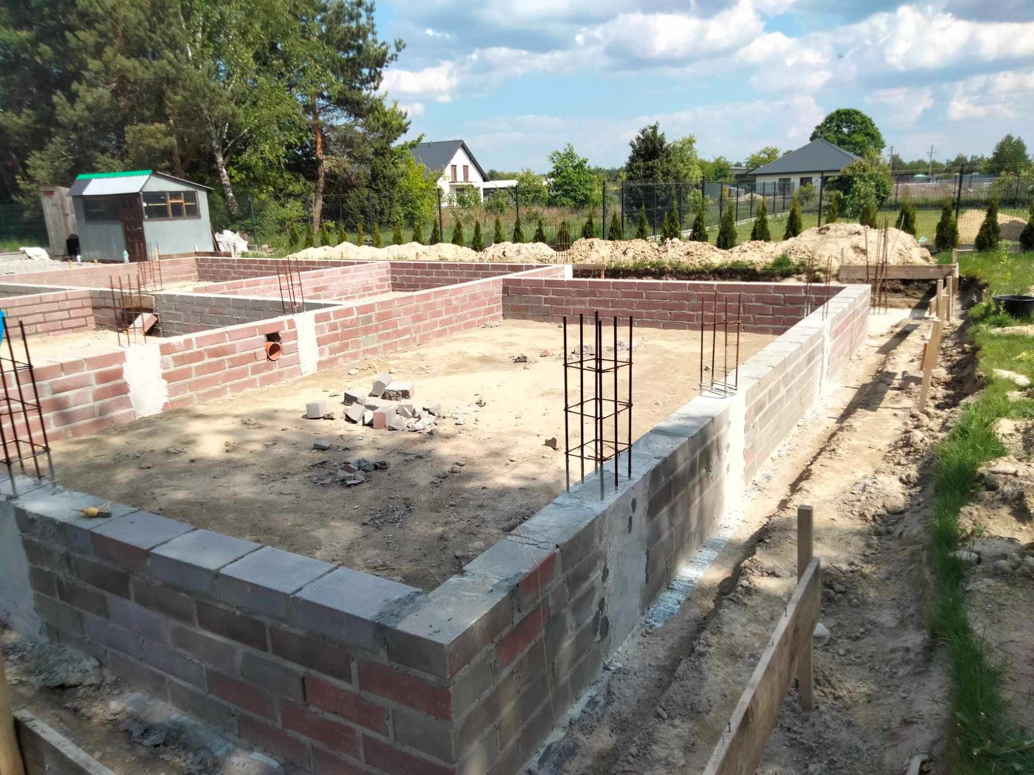 Ceglany fundament domu w trakcie budowy, zbrojenie pionowe, widok na działkę z domami w tle, słoneczny dzień.
