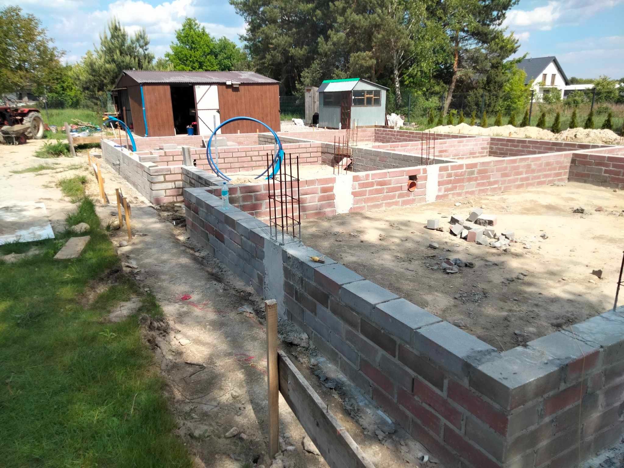 Wznoszenie fundamentów z cegły i bloczków betonowych. Widoczne zbrojenia, szalunki i przygotowane instalacje. W tle budynki gospodarcze i drzewa.