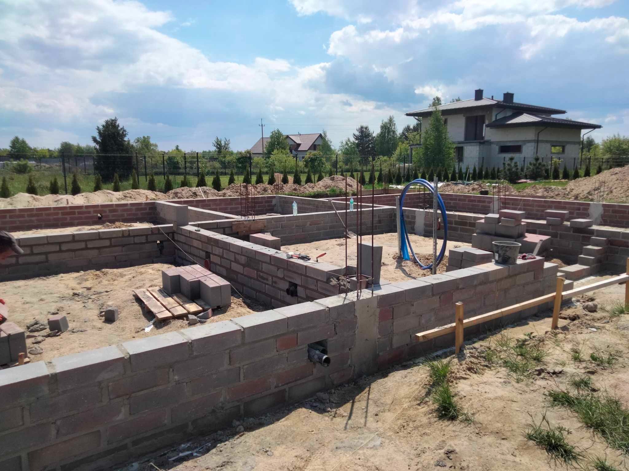 Murowane ściany fundamentowe z czerwonej cegły na tle domu w budowie i błękitnego nieba z chmurami. Widoczne zbrojenia i instalacje. Etap budowy.