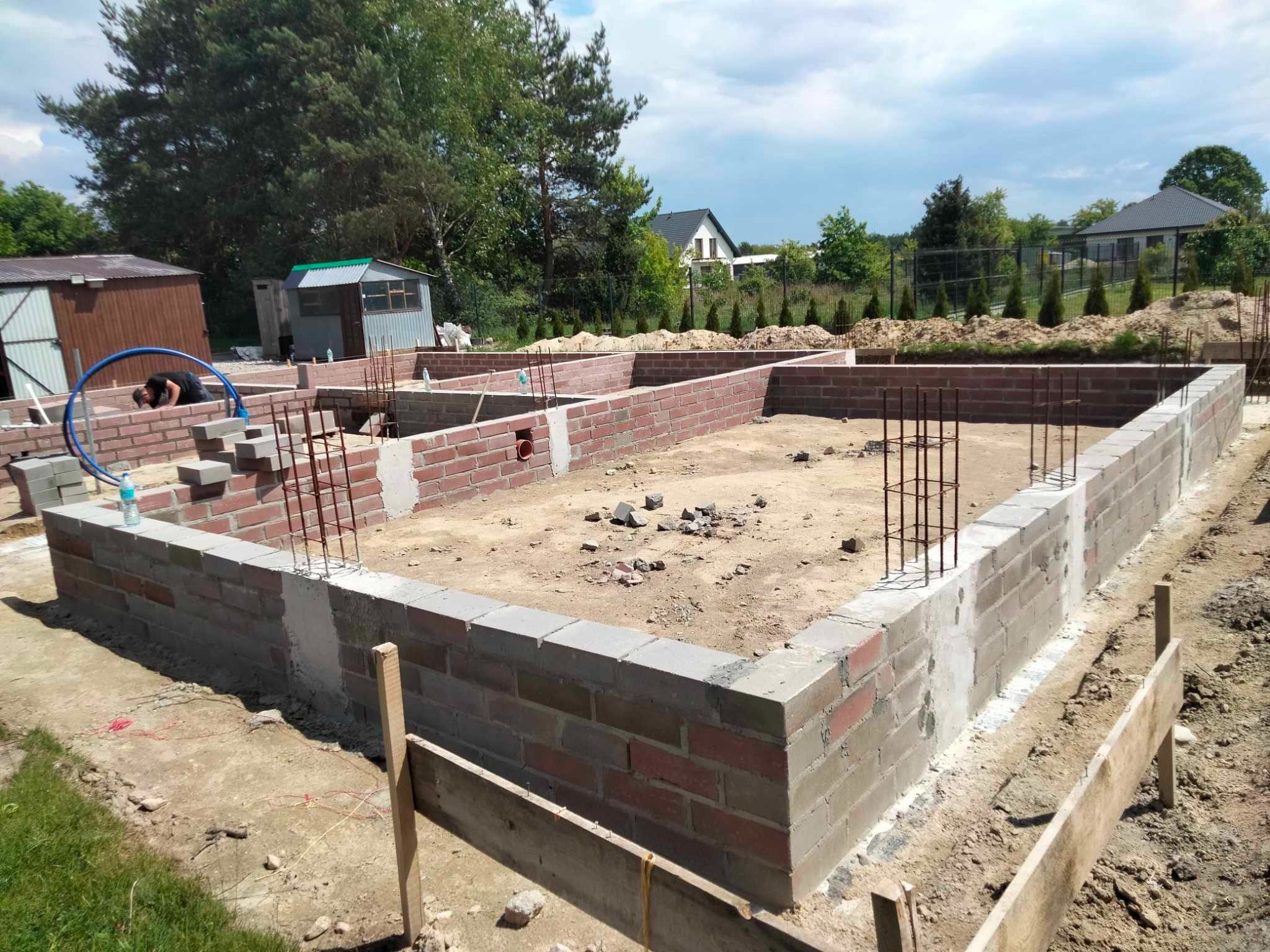Wykopane i wymurowane fundamenty domu z czerwonej cegły i szarego betonu. Widoczne zbrojenia, teren budowy i częściowo pracownik. W tle zieleń i budynki.