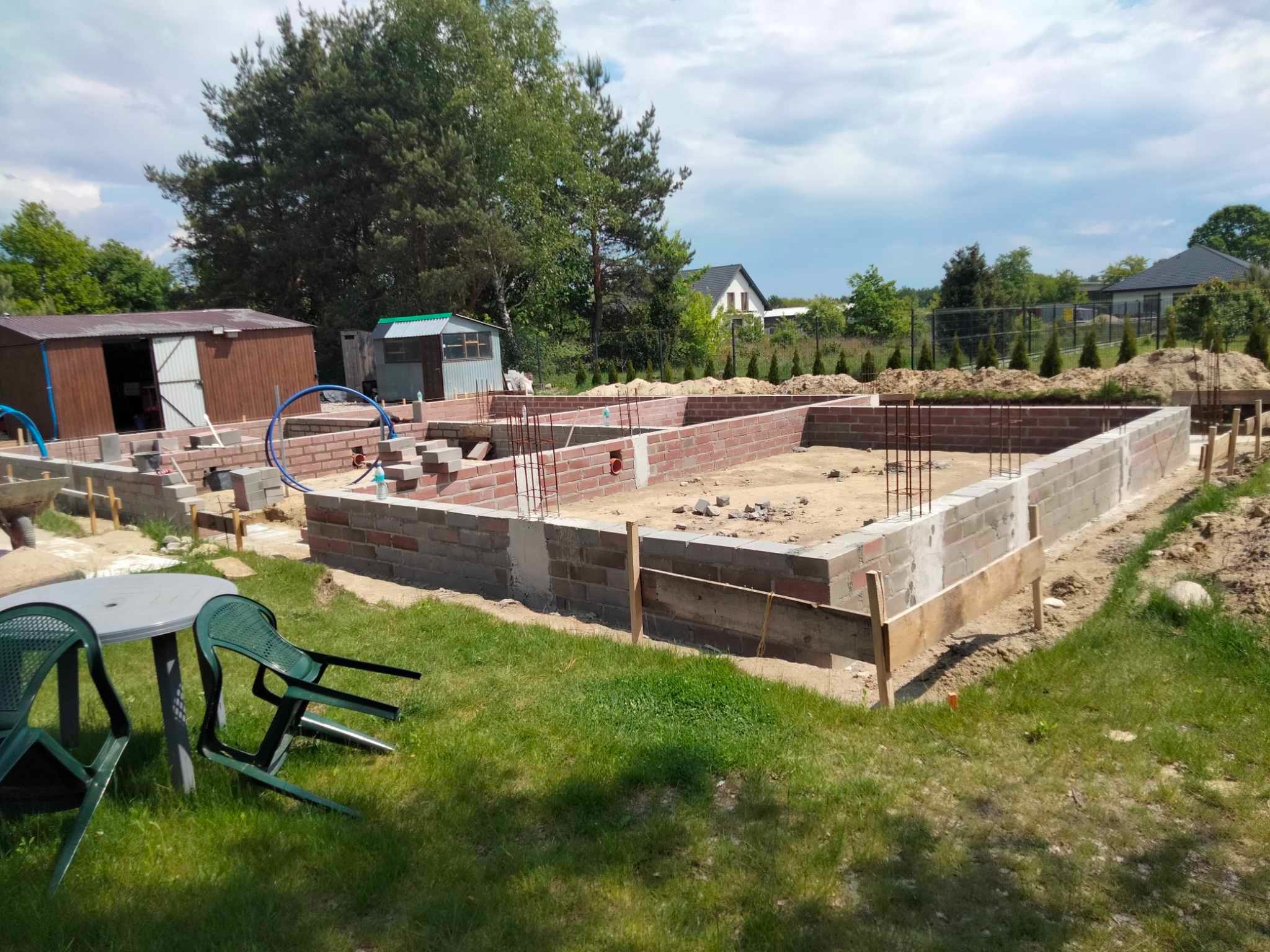Fundamenty domu w trakcie budowy: murowane ściany z betonu i cegły, wystające pręty zbrojeniowe, widoczne szalunki. W tle zieleń i zabudowania.