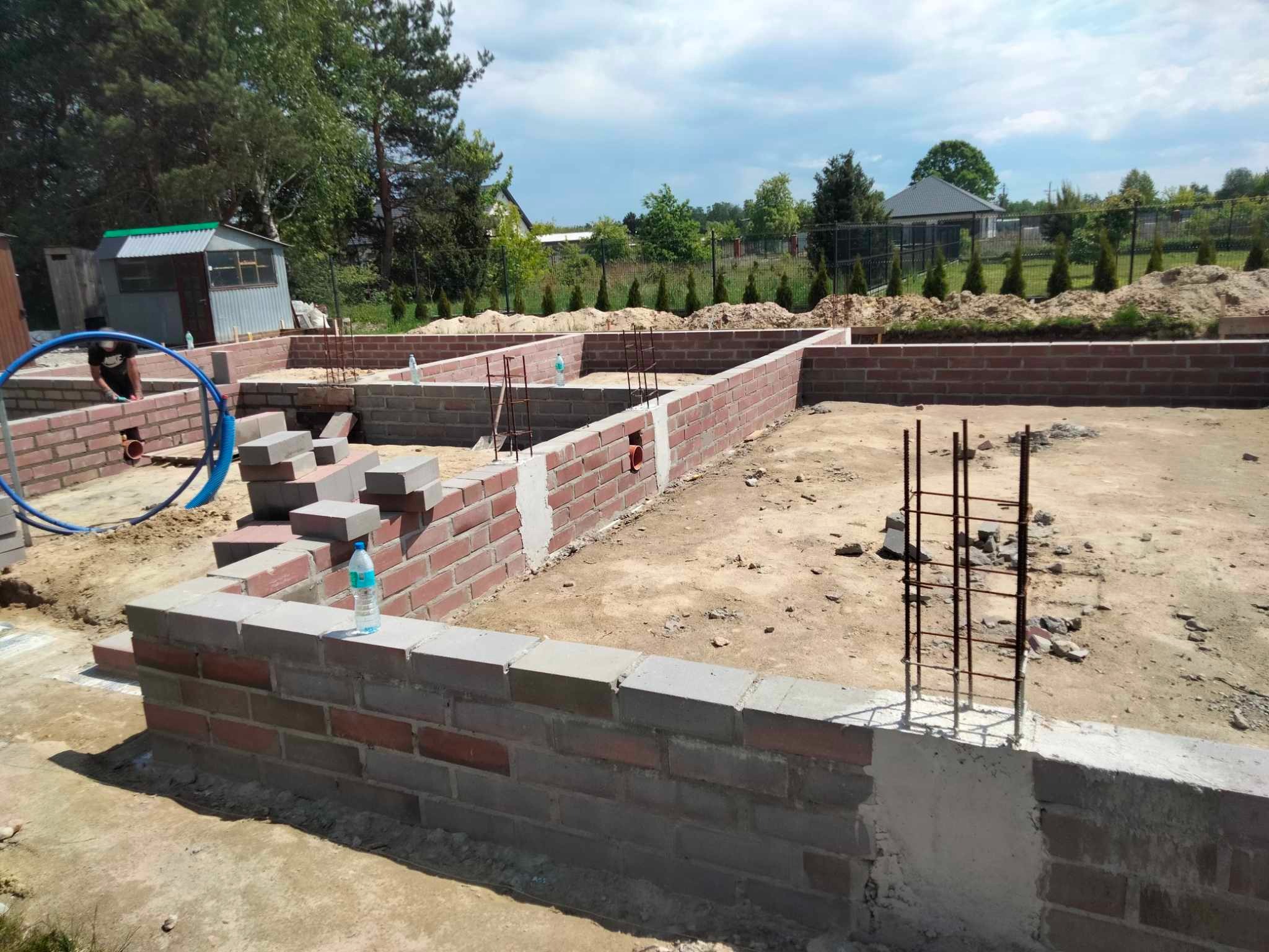 Ceglane fundamenty domu w trakcie budowy, z wystającymi prętami zbrojeniowymi i butelką wody. Widoczny pracownik i niebieski wąż. Tło: zieleń i zabudowania.