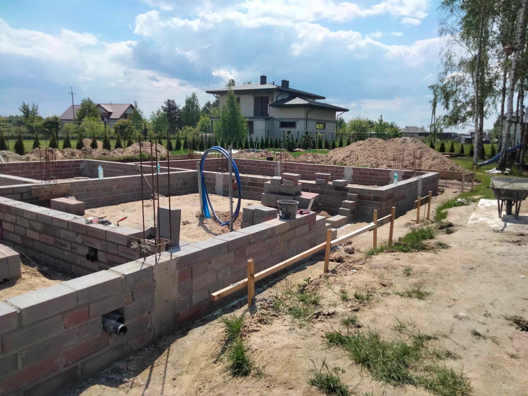 Budowa domu jednorodzinnego: wznoszone mury fundamentowe z pustaków, widoczne zbrojenia, rury instalacyjne i przygotowany teren pod dalsze prace. Dom w tle.