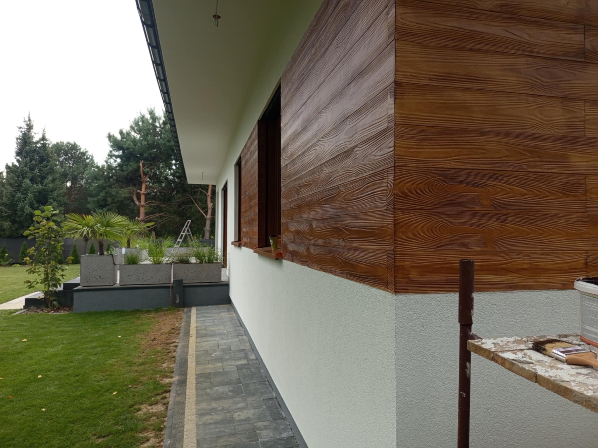 Elewacja domu z efektem drewna, widoczna z boku, z fragmentem chodnika i ogrodu. Na pierwszym planie rusztowanie z pędzlem i puszką farby.