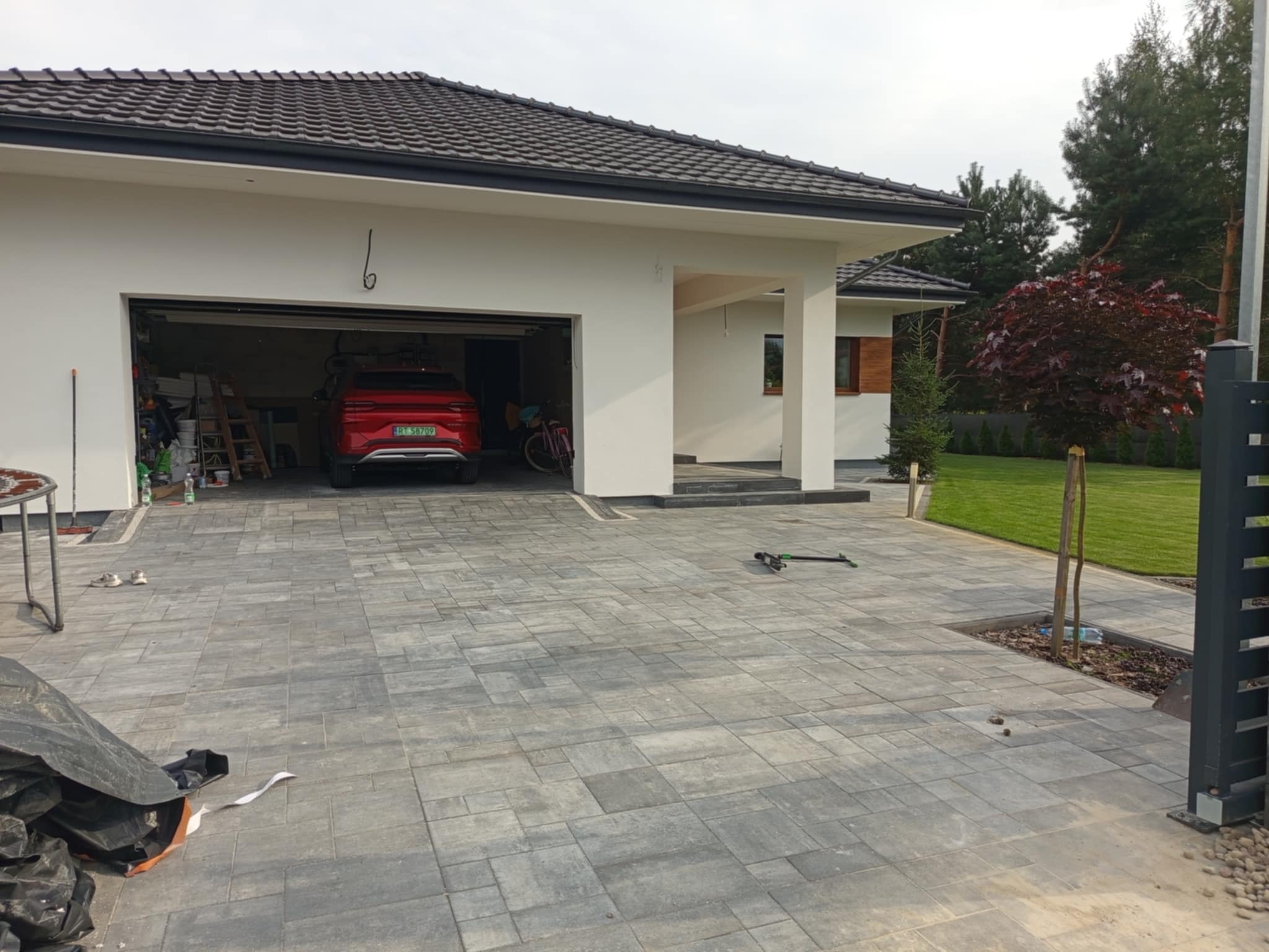 Ułożona szara kostka brukowa na podjeździe przed nowoczesnym domem z otwartym garażem, w którym stoi czerwony samochód. Widoczny trawnik i drzewa w tle.