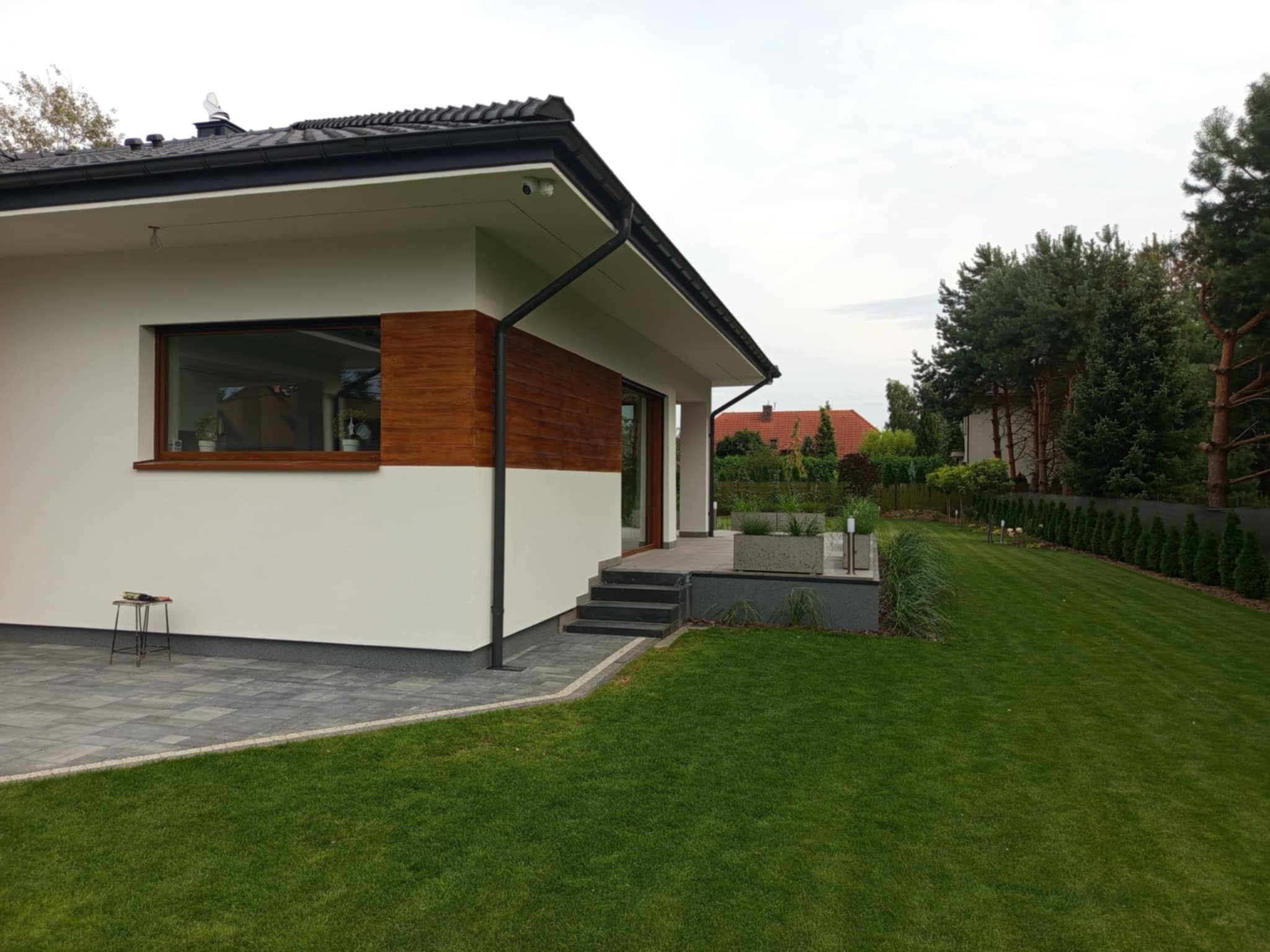 Nowoczesny dom jednorodzinny z białymi ścianami, drewnianymi akcentami i szarą kostką brukową. Widoczny zadbany trawnik i elementy architektury krajobrazu.