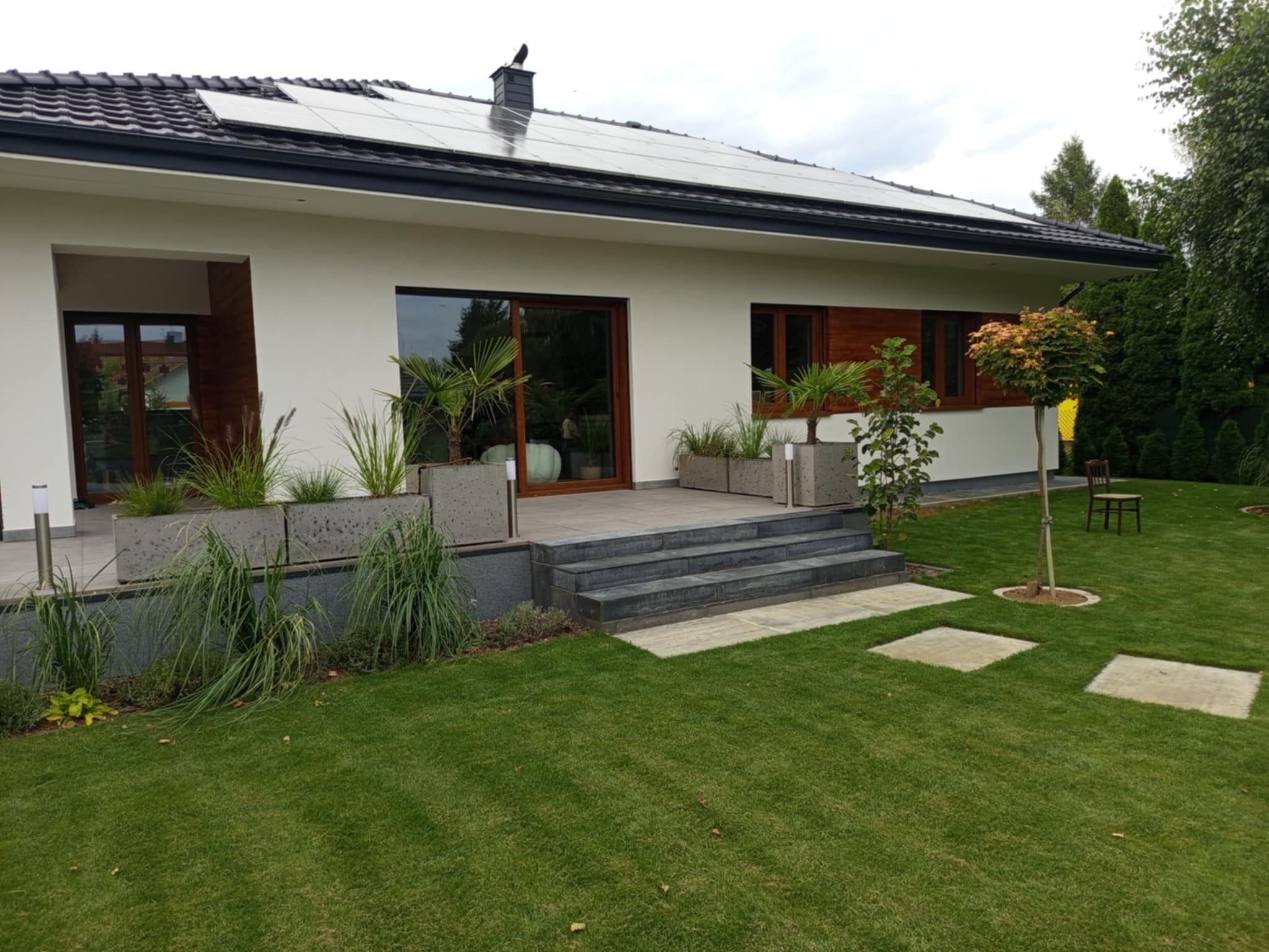 Nowoczesny dom jednorodzinny z panelami słonecznymi na dachu, tarasem z roślinami w donicach i zadbanym trawnikiem z elementami z kostki.