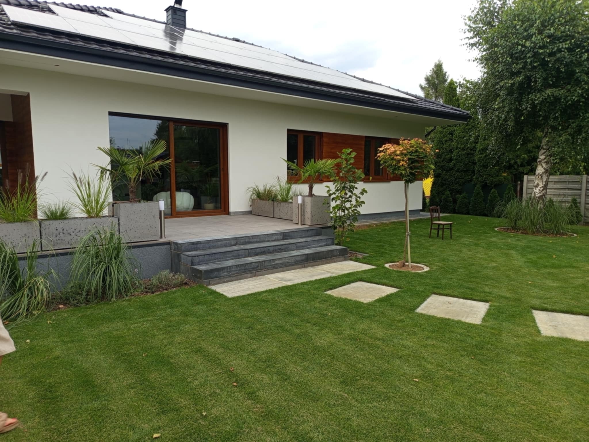 Nowoczesny dom jednorodzinny z białymi ścianami, panelami słonecznymi na dachu i zadbanym trawnikiem z kamiennymi płytami. Widoczne rośliny w betonowych donicach.