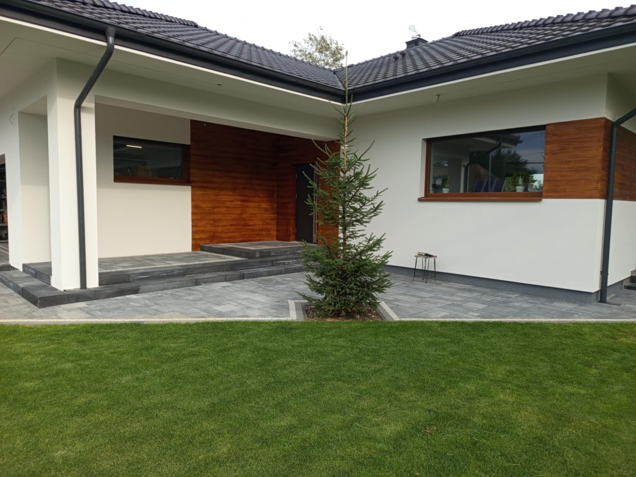 Nowoczesny dom z jasną elewacją, drewnianymi akcentami i szarą kostką brukową. Na pierwszym planie trawnik i małe drzewko iglaste. Minimalistyczny styl.