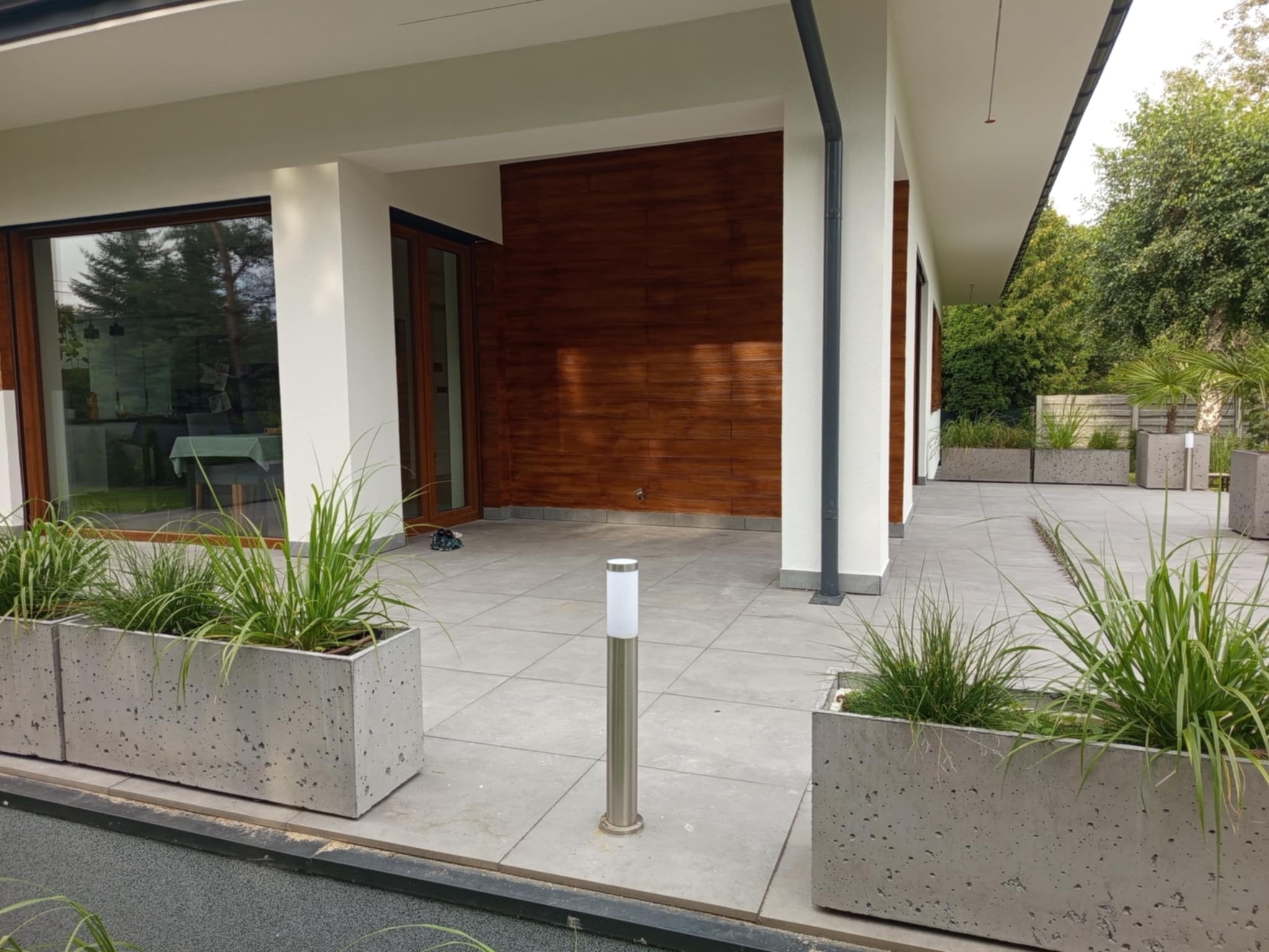 Nowoczesny taras z szarych płyt, betonowe donice z zielenią, oświetlenie punktowe. Elewacja z drewnianymi panelami i białymi kolumnami. Minimalistyczny design.
