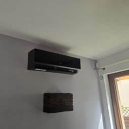 Power Sun Polska - Czarna klimatyzacja TCL na ścianie w mieszkaniu w Krakowie. Widoczny drewniany element dekoracyjny pod klimatyzatorem i okno z widokiem na zewnątrz.