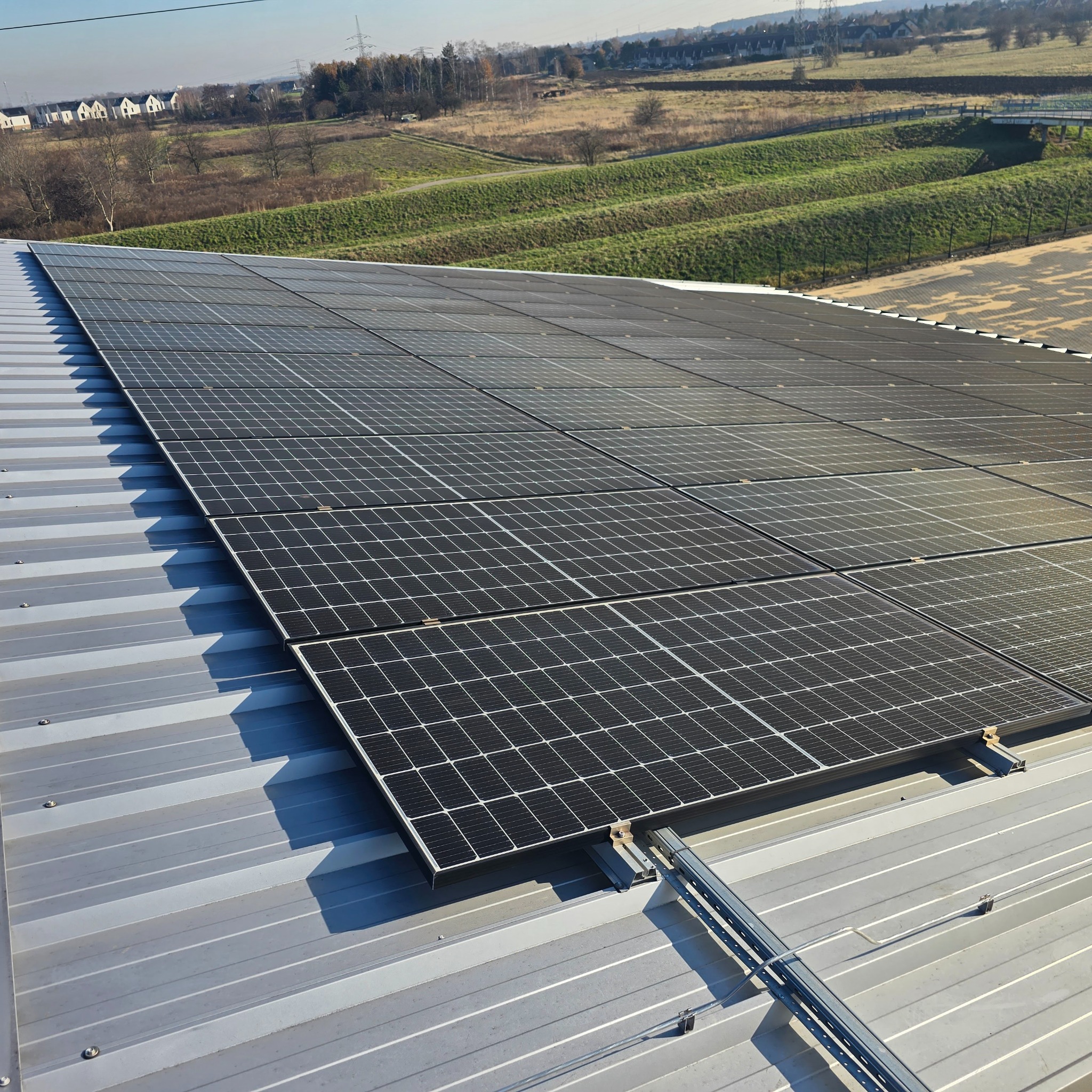 Rząd paneli fotowoltaicznych zamontowanych na metalowym dachu, z widokiem na zielone wzgórza i zabudowania w tle.